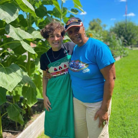 Board Members Margo Wright and Kris De la Torre share a common passion for gardening, smiling in front of tall vegetables in grow beds