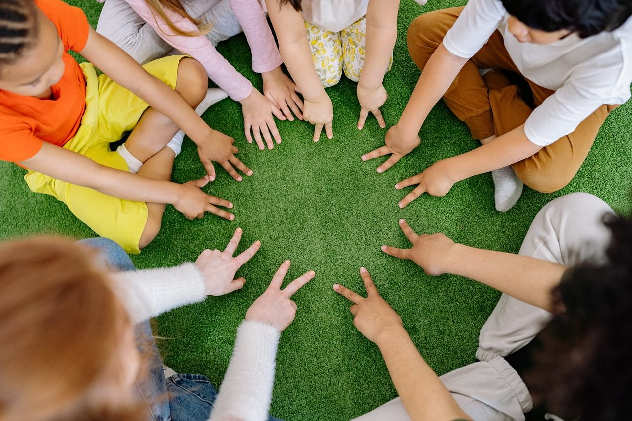 A circle of children from all backgrounds and ethnicities form peace signs with their hands.