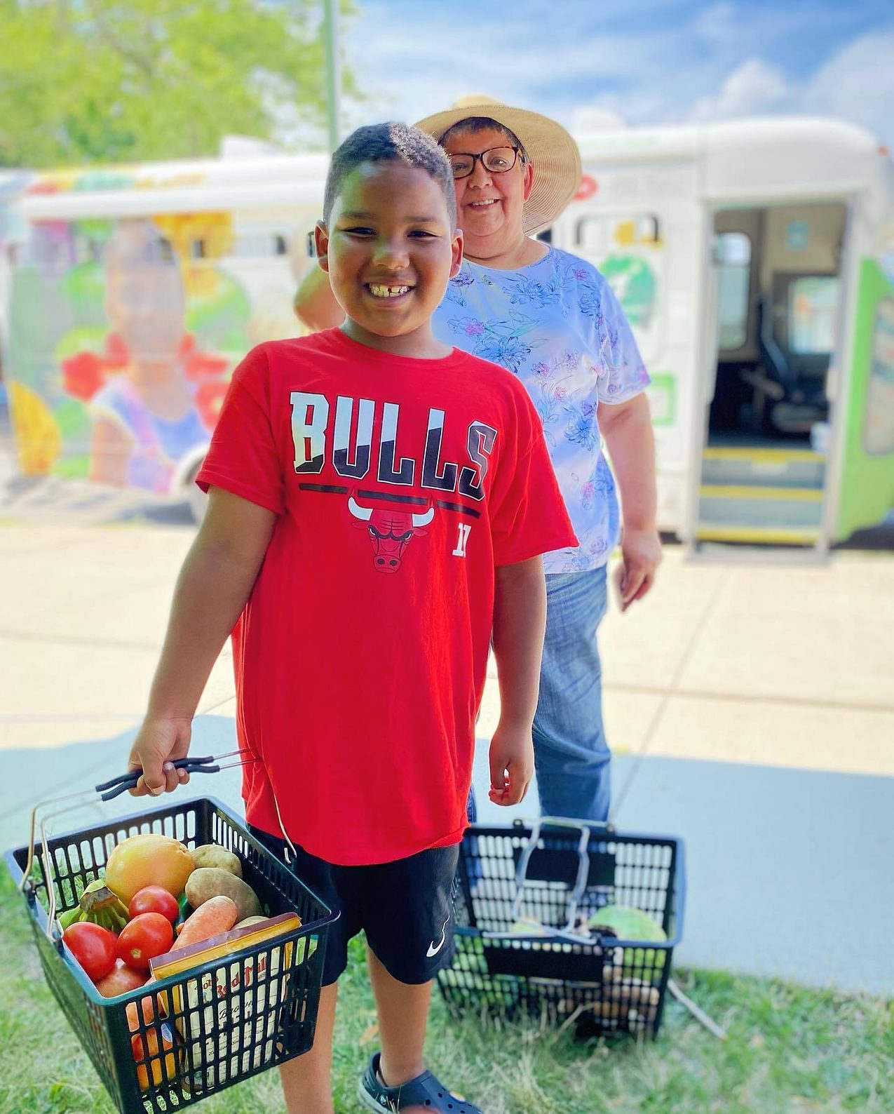 A smiling boy wearing a Chicago Bulls shirt carries a shopping basket full of vibrant fresh produce